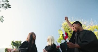Mezuniyet elbisesi giyen, kep takan ve güneşli bir günde kutlama yapan mutlu, çok ırklı, beyaz ve Hintli erkek ve kadın arkadaşlar grubu. Kutlama, mezuniyet, eğitim ve arkadaşlık kavramı.