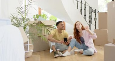 Mutlu adam ve partneri yeni eve taşınıyorlar, yerde oturuyorlar yeni evlerinin olduğu kutularla telefon kullanıyorlar, ev sahipleri, ipotekler. Yeni başlangıçlar, aile hayatı, mutluluk, yaşam tarzı ve ilişki kavramı.