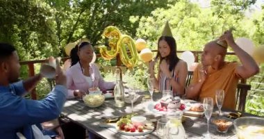 Bir grup mutlu çok ırklı ve siyah erkek ve kadın arkadaş güneşli bir günde bahçede bir doğum günü partisinde kadeh kaldırıyorlar. Kutlama, doğum günü, parti, yaz ve arkadaşlık kavramı.