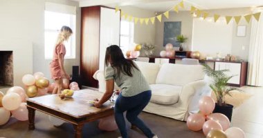 İki mutlu çok ırklı ve beyaz bayan arkadaş güneşli oturma odasında bir doğum günü partisi için masa ve dekorasyon hazırlıyor. Kutlama, doğum günü, parti, yaz ve arkadaşlık kavramı.