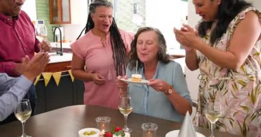 Doğum günü pastasında mum üfleyen ve çeşitli son sınıf arkadaşlarıyla kadeh kaldıran mutlu son sınıf kadını. Kutlama, doğum günü partisi, son sınıf yaşam tarzı ve arkadaşlık konsepti.