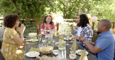 Bir grup mutlu, çok ırklı, siyah ve Çinli erkek ve kadın arkadaş mum üflüyor ve terasta bir doğum günü partisinde kadeh kaldırıyorlar. Kutlama, doğum günü, parti, yaz ve arkadaşlık kavramı.