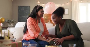 İki mutlu çok ırklı ve siyah bayan arkadaş kanepeye oturup güneşli oturma odasındaki bir doğum günü partisinde sohbet ediyorlar. Kutlama, doğum günü, parti, yaz ve arkadaşlık kavramı.