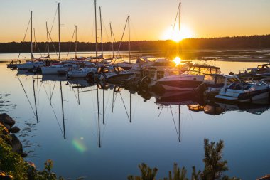 Körfezdeki rıhtıma demirlemiş çok sayıda yat, tekne ve gemi var. Beyaz yatlar parlak turuncu günbatımına ve ufuktaki ormana karşı..