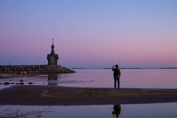 A man takes pictures of the church against the sea. The church in the background of the lake. A tourist takes pictures of the attraction. Beautiful pink sunset.