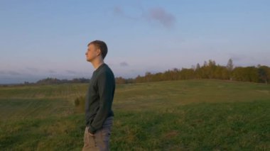 Bir adam elleri ceplerinde durur ve ormanın üzerindeki güzel günbatımını seyreder. Doğanın ve günbatımının güzelliğini düşünüyorum. Büyük yeşil bir çayır, etrafında bir sürü yeşil alan var..