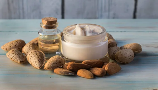 Facial cream and almond oil surrounded by natural almonds, on a white wooden board