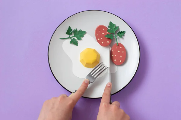 Hands with fork and knife cut paper fried eggs with sausage. Realistic handmade paper objects. Paper art and craft. Minimal art food concept