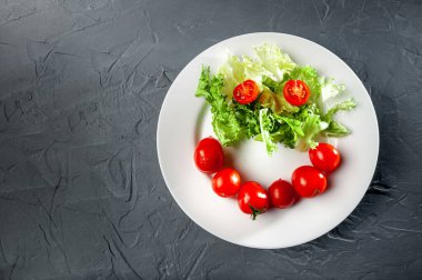 Yeşil salata ve kiraz domatesleri gri arka planda beyaz bir tabakta emoji formunda ve biçiminde.