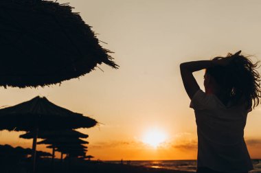 Gün batımında genç bir kız sahilin yanındaki şemsiyelere, siletlere bakar.