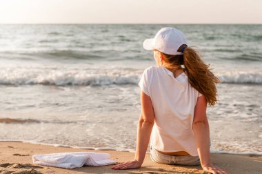 Sporcu koşudan sonra sahilde oturur ve gün batımını seyreder. Beyaz tişörtlü ve şapkalı kız.