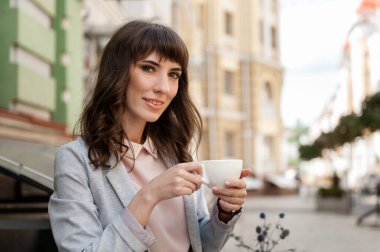 Elinde kahve olan bir kız bir kafede oturur ve kameraya bakar.