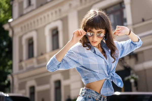 Outdoor portrait of yong beautiful happy smiling woman wearing stylish sunglasses, blue jeans blouse, blue mom jeans, with small quilted bag. Model posin in street of european city. Copy space — Stock Photo, Image