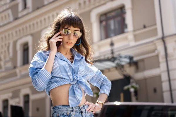 Outdoor portrait of yong beautiful happy smiling woman wearing stylish sunglasses, blue jeans blouse, blue mom jeans, with small quilted bag. Model posin in street of european city. Copy space — Stock Photo, Image