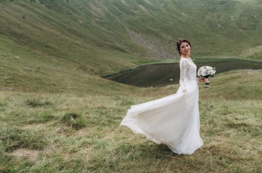 Gelin gölün yanındaki dağlarda. Gelin uzun bir gelinlik giyiyor, dantelli ve arkası açık.