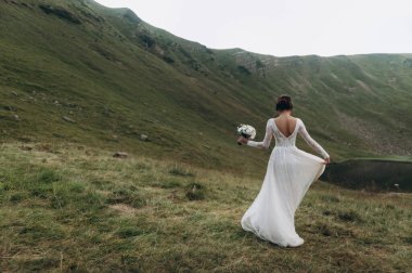 Gelin gölün yanındaki dağlarda. Gelin uzun bir gelinlik giyiyor, dantelli ve arkası açık.