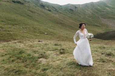 Gelin gölün yanındaki dağlarda. Gelin uzun bir gelinlik giyiyor, dantelli ve arkası açık.