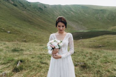 Elinde bir buket çiçekle gelin dağlardaki göle karşı duruyor.