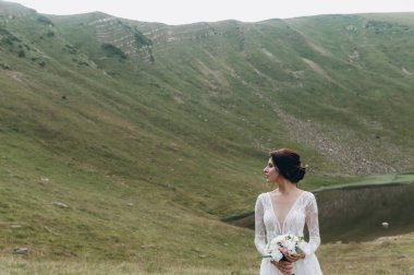 Gelin gölün yanındaki dağlarda. Gelin uzun bir gelinlik giyiyor, dantelli ve arkası açık.