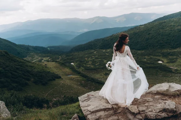 the bride stands on a rock in the mountains. Wind develops white bride dress