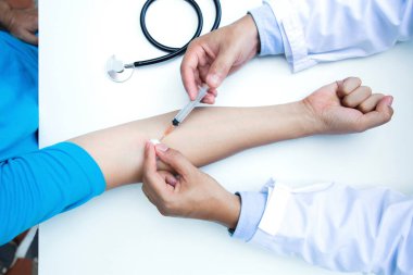 Hastane odasında steteskoplu ve şırıngalı bir doktor var. Doktor hasta hastanın koluna şırınga enjekte ediyor. Üst görünüm.