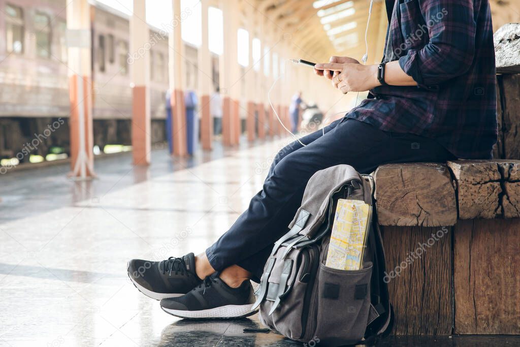 Young man traveler sitting with using mobile phone choose where to travel and bag waiting for train at train station.backpacker at the train station and looking on mobile phone for plan to travel