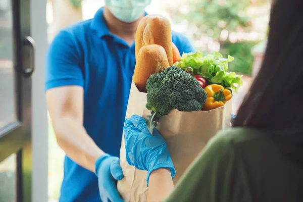 Yüzüne maske takan ve mavi üniformalı hijyen eldivenleri giyen teslimatçı evin önündeki kadın müşteriye yiyecek, meyve ve sebze dolu bir çanta veriyor. Covid19.sırasındaki teslimat servisi. Yeni normal