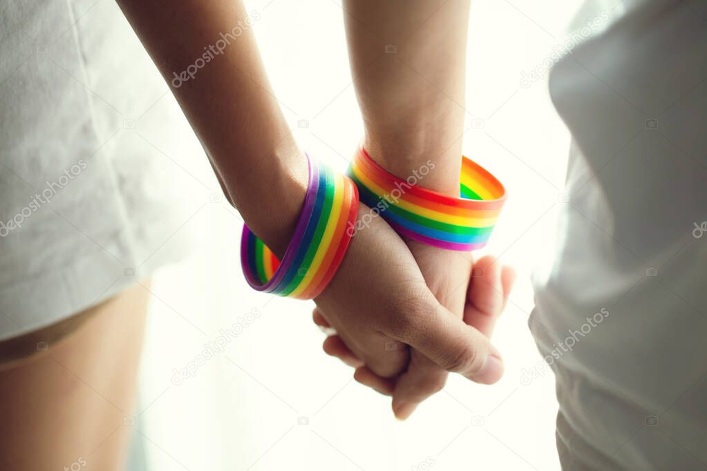 Closeup lesbian couple holding hands with a rainbow-patterned wristban on their wrists.Concept of LGBT, activism, community and freedom.