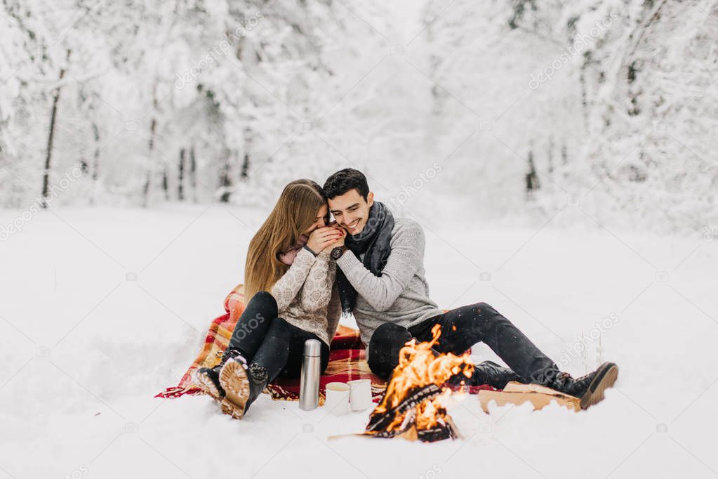 Young couple on a date in the winter forest is heated by the fire and drink cocoa