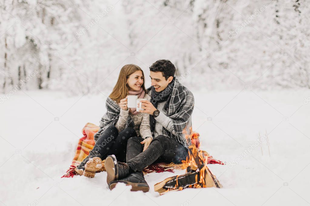 Young couple on a date in the winter forest is heated by the fire and drink cocoa