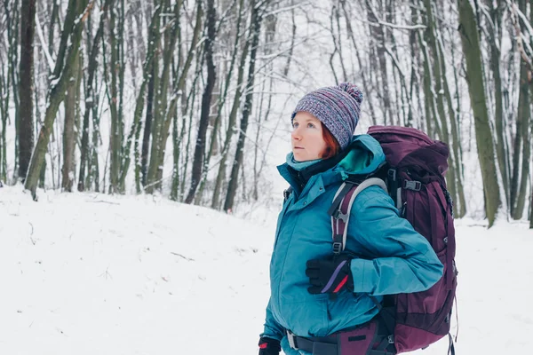 Chica joven con mochila mira hacia arriba senderismo a través del bosque de invierno. Mira desde el lado izquierdo Imágenes de stock libres de derechos