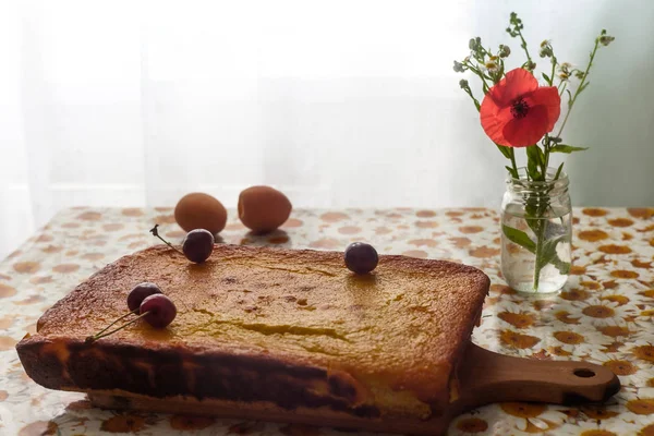 Homemade cheesecake on a wooden slicing board, flowers in the bottle, apricots and sherries on the table. View from the side — Stock Photo, Image