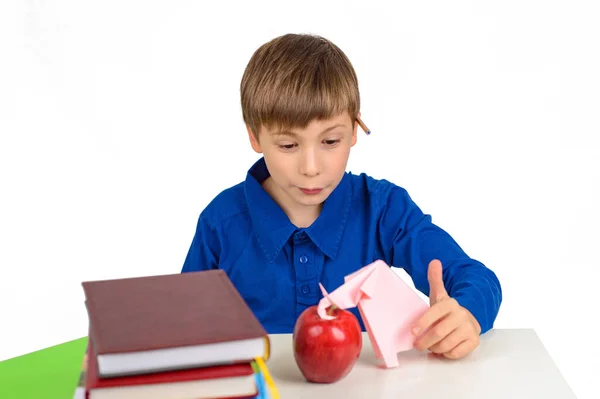 child development, origami: a schoolboy boy in a blue T-shirt sits at the table and plays with an elephant from paper, next to a stack of textbooks and a red apple