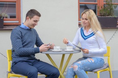 Genç bir adam ve uzun saçlı sarışın bir kız kahve ya da çay içerler masadaki bir kafenin yanında, bir adam bir kıza telefonundan bir şey gösterir, gülerler.