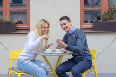 Genç bir adam ve uzun saçlı sarışın bir kız kahve ya da çay içerler masadaki bir kafenin yanında, bir adam bir kıza telefonundan bir şey gösterir, gülerler.