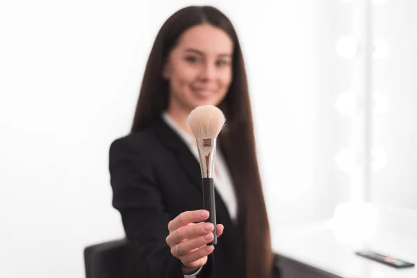 girl, makeup artist, with long dark hair in a business suit holding a powder brush on an outstretched hand, on a white background