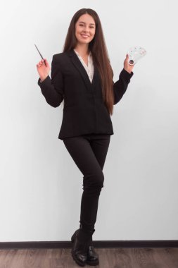 Makyajlı, uzun siyah saçlı, iş elbisesi içinde elinde bir fırça ve beyaz arka planda kremleri karıştırmak için palet tutan bir kız.