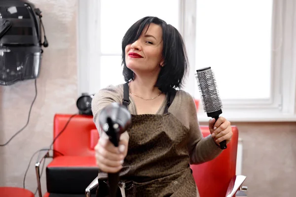 a black haired woman hairdresser sits in a hairdressers chair and laughs as she holds a brushing and hairdryer