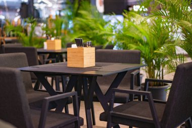 Kahverengi masaları ve sandalyeleri olan kafe, malzeme masalarında sofra takımı, yeşil palmiye ağaçları etrafında