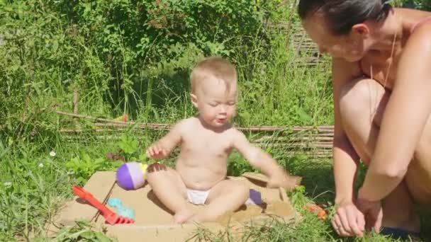 小さな赤ちゃんの女の子はサンドボックスに砂の上に座って 緑の庭の背景にカラフルなおもちゃで遊ぶ — ストック動画