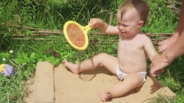 Anne, küçük kızına koruyucu güneş kremi sürüyor. Kum havuzunda renkli oyuncaklarla oynayan tatlı bir bebek.