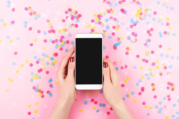 A smartphone in woman hands with perfect manicure. Colorful confetti background. The screen is a mock up, easy to use for any design. Copy space. Flat-lay, top view.