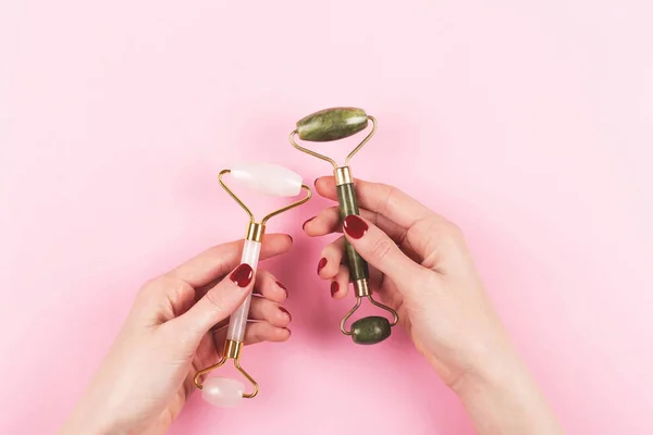 Two jade rollers in woman hands on pink background.