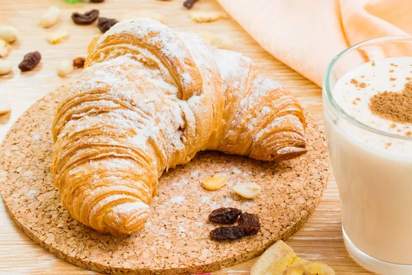 Croissant in powdered sugar and glass of milk wiht heart from cinnamon on a wooden table