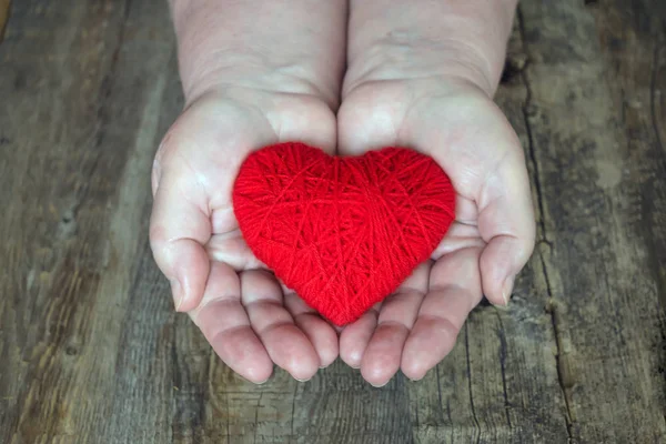 Organ donation, help someone. Adult woman hands giving thread red heart. Hands of adult woman, mother holding a red heart in the palm on dark wooden background