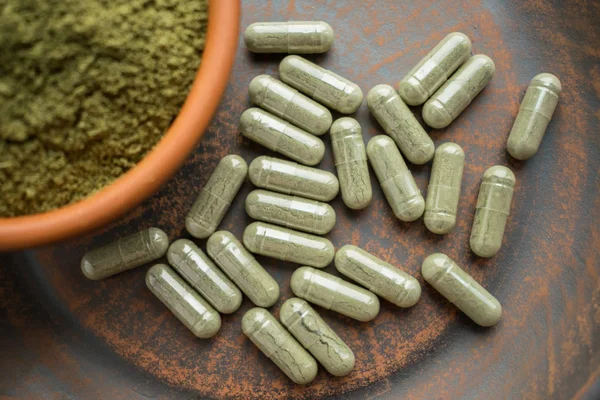 Kratom yeşil kapsül ve toz kahverengi plaka üzerinde ek. Bitkisel ürün alt-tıp kratom opioid. Ev alternatif ağrı ilaç, opioid bağımlılığı, tehlikeli ağrı kesici, aşırı doz. Yakın çekim. Seçici odak