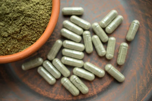 Kratom yeşil kapsül ve toz kahverengi plaka üzerinde ek. Bitkisel ürün alt-tıp kratom opioid. Ev alternatif ağrı ilaç, opioid bağımlılığı, tehlikeli ağrı kesici, aşırı doz. Yakın çekim. Seçici odak