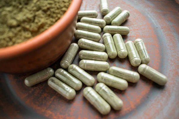 Kratom yeşil kapsül ve toz kahverengi plaka üzerinde ek. Bitkisel ürün alt-tıp kratom opioid. Ev alternatif ağrı ilaç, opioid bağımlılığı, tehlikeli ağrı kesici, aşırı doz. Yakın çekim. Seçici odak