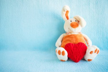 Sevimli yumuşak oyuncak tavşan ve mavi bir arka plan üzerinde kırmızı örgü kalp. Tüylü şirin tavşan kalp ile oturan koca ayaklı. Aşk, Sevgililer günü, romantizm, hassasiyet kavramı. Kopya alanı