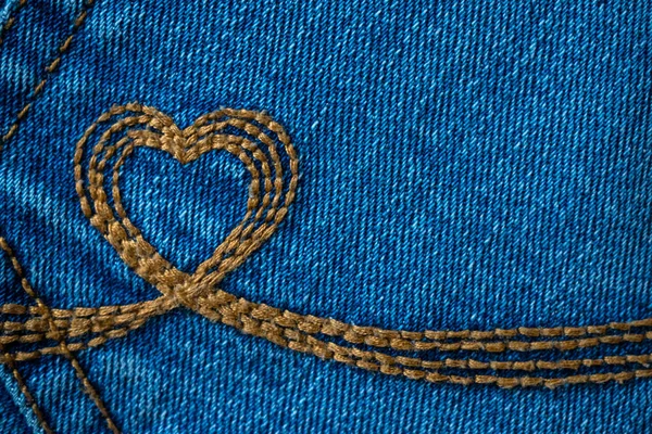 Embroidery in the shape of a heart on a denim pockets. Blue denim background with brown stitch